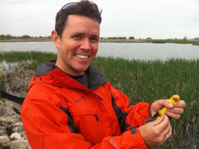 Researcher Jeb Owen holds a yellow Tanager bird in his hands. He is wearing a red raincoat, smiling at the camera, standing in a marshy area in front of a body of water.